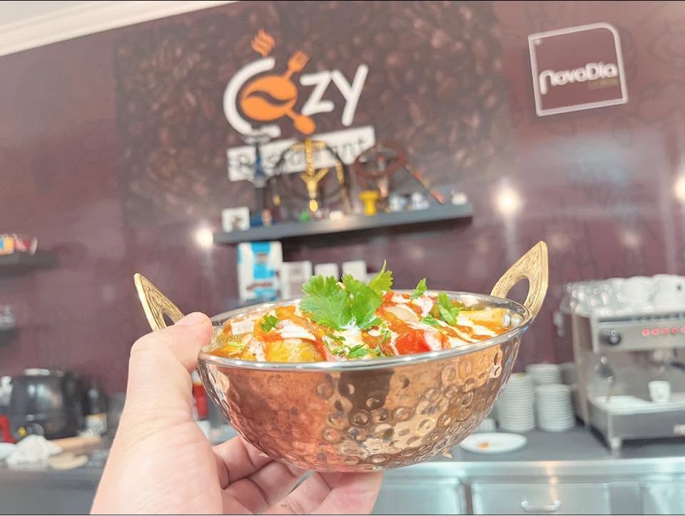 A copper karahi of creamy curry topped with fresh coriander, held in front of the Cozy Restaurant wall logo